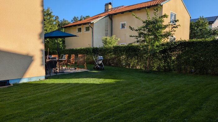 Trädgård med gräsmatta, trädgårdsmöbler under parasoll, en barnvagn och ett gult hus i bakgrunden.