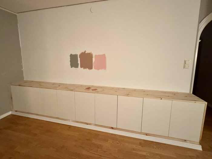 Inbyggd bokhylla under uppbyggnad med fem vita dörrar och furu toppskiva; tre färgprover målade på väggen ovanför.