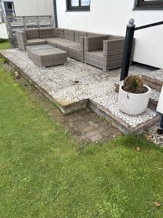 Utomhusplats med mosaikplatta och rottingmöbler, intill gräsmatta. Trappräcke och kruka syns, förbereder för uterum.
