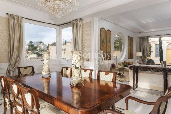 Ljust vardagsrum med matbord, stolar, och stora fönster som visar utsikt över staden. Elegant inredning med kristallkrona och dekorativa vaser.