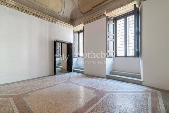 Tomt rum med mönstrat stengolv, högt i tak, stora fönster med trädetaljer och en dörröppning till vänster.