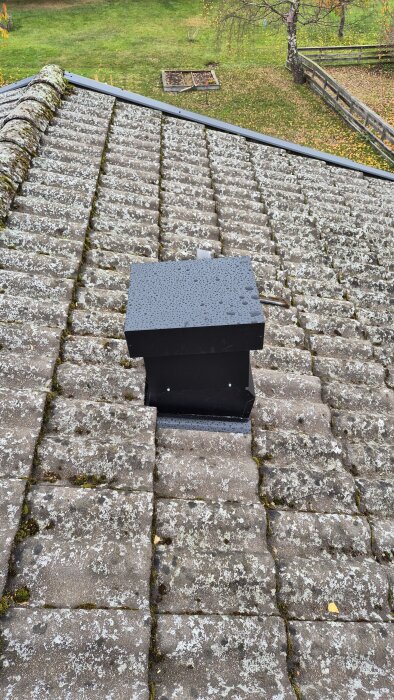 Svart fläktkåpa installerad på tak med mossa och lav på tegelpannor, del av renoveringsprojekt.