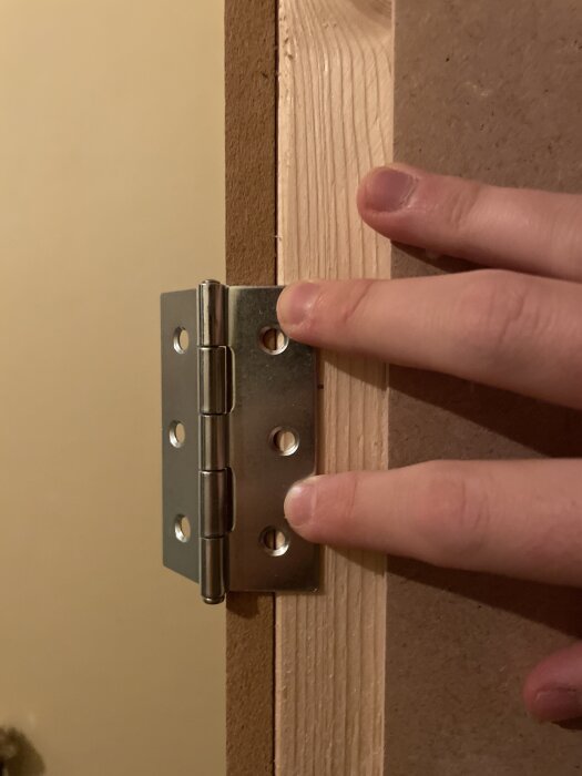 Skinnende metalhængsel holdes mod en træramme med MDF-plade. Fingrene viser først mulig skrueplacering til garderobedør.