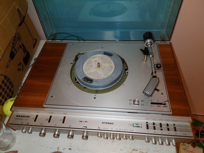 Gammal Skantic Harmoni 17S vinylspelare utan skiva, på ett bord med träyta och en kartong med borrbild i bakgrunden.