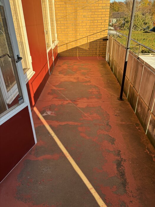 Balkong i radhus med utsikt över trädgård, ytan har flera synliga sprickor och flagnande färg, omgivet av röd vägg och tegel.