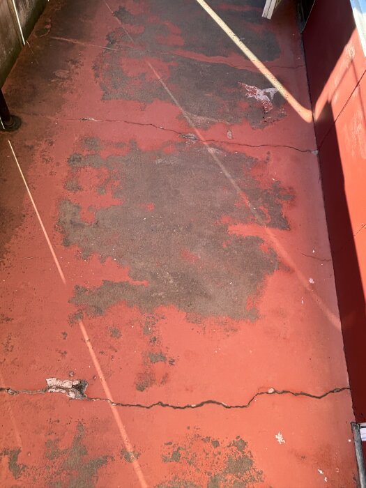 Röd betongbalkong med synliga sprickor och flagnande färg. Balkongen är ovanpå kök och vardagsrum, och behöver repareras inför vintern.