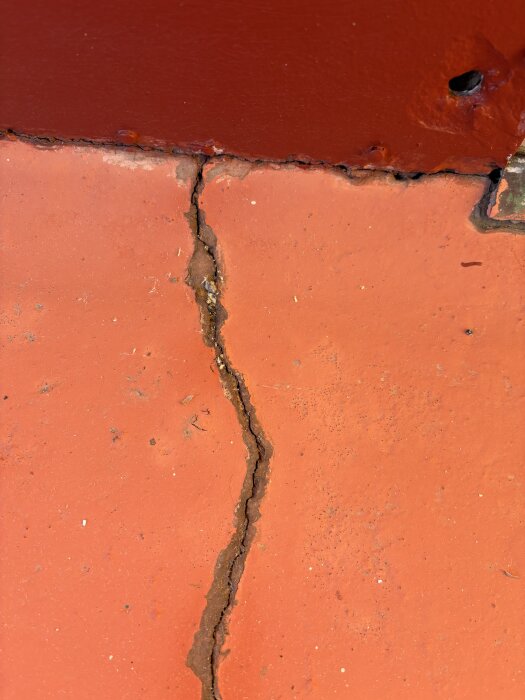 Spricka i balkongens betongyta från närbild, visar gammal yta med röda inslag som behöver repareras inför vintern för att förhindra fuktinträngning.