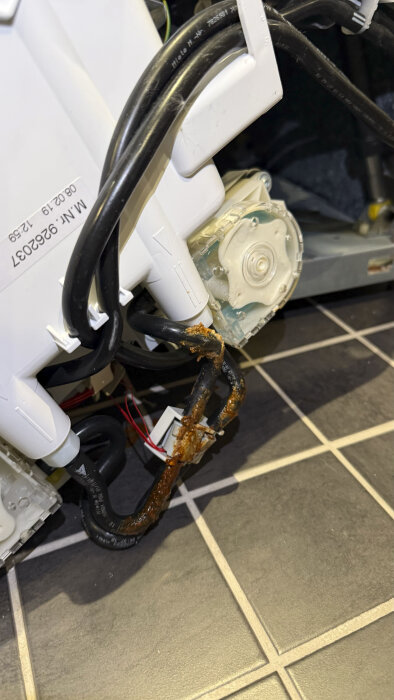 Inre delen av en tvättmaskin med rostiga och korroderade slangar samt elledningar, visar tecken på läckage och skador på plastdetaljer.