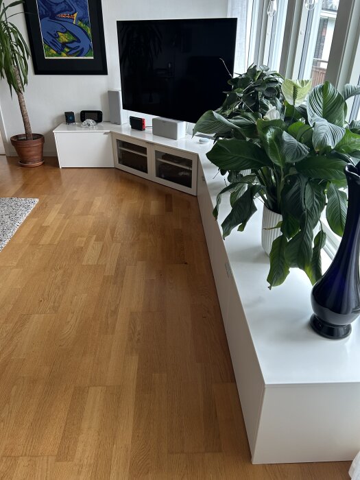 Vit kombinerad tv- och fönsterbänk med tv, växter och ljudutrustning framför stort fönster. Möbeln består av Ikea Bestå-enheter.