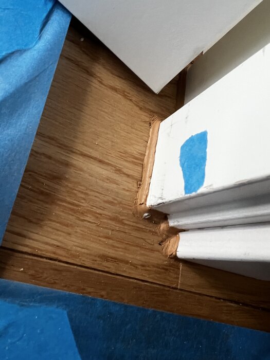 Vit dörrkarm mot trägolv med snickeridetalj i hörn. Tejp syns nära kanten.