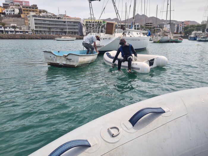 Två personer i små båtar på vattnet nära hamnbyggnader och segelbåtar.