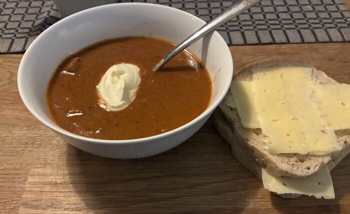 En skål med Felix Gulashsoppa toppad med en klick grädde och en sked, bredvid en macka med skivad ost på ett bord.