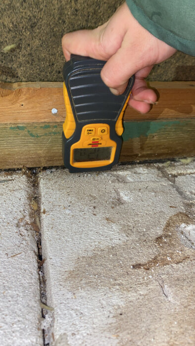 En hand håller en fuktmätare mot en trälist nära ett betonggolv.