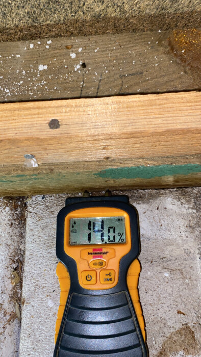 Fuktmätare visar 14% på träplanka nära golvet, omgivet av betong och trä.