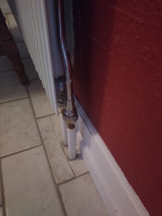 Radiatoranslutning med rör och kopplingar mot röd vägg och ljus golvplatta.