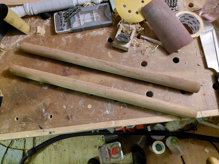 Två korta rundstavar av trä, cirka 33 mm i diameter och 570 mm långa, ligger på en arbetsbänk med verktyg runtomkring.