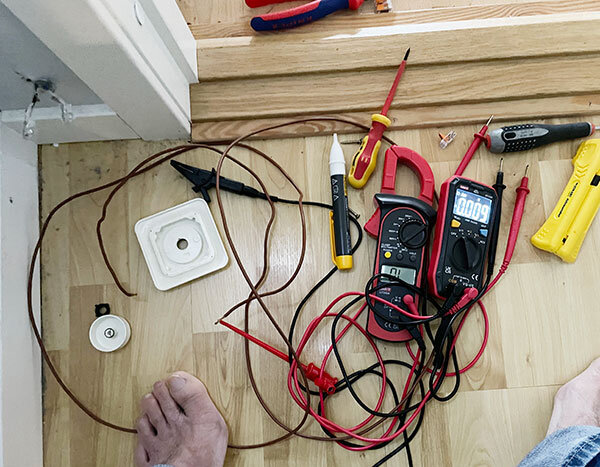 Multimeter, sladdar och elektriska verktyg ligger på golvet nära ett uttag, med en fot synlig i hörnet av bilden.