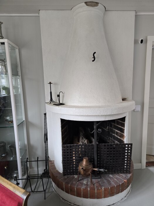 Vit putsad eldstad med sotlucka och svart galler framför, ståendes på tegelbas. Eldstädet är dekorerat med en kopparkokare.