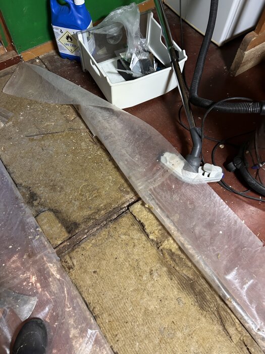 Bild på ett öppet golv i en snickarbod med synliga, murkna reglar och blöt gul isolering. Plastöverdrag och verktyg synliga i rummet.