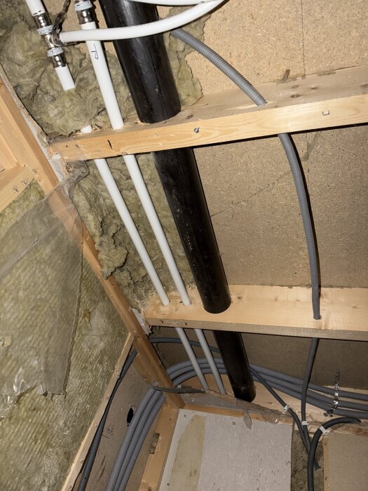 Svart gjutjärnsrör bredvid vita och grå elkablar genom trätak, omgiven av isolering, i ett hus från 1976.