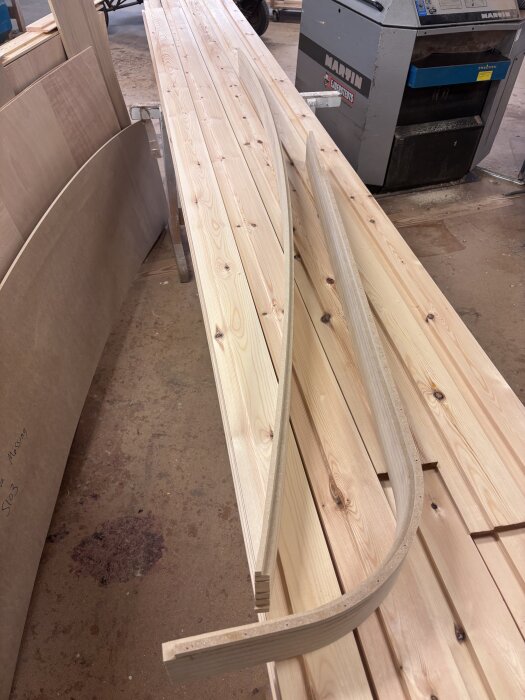 Sammanlimmade och frästa trästycken i lager för svängda socklar på verkstadsgolv, med en träbearbetningsmaskin i bakgrunden.