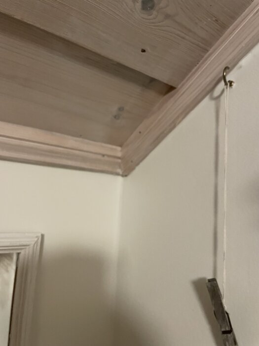 Vinklat takhörn av trä med ljus list och krok för snöre mot vit vägg.