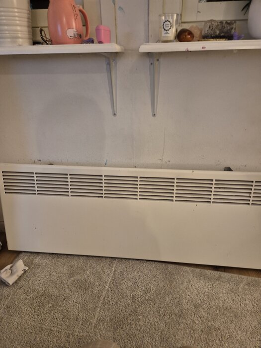 Väggmonterad vit radiator under hyllor med olika föremål.