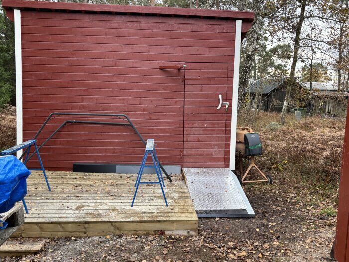 Röd byggnad med nybyggd ramp av metall bredvid en träplattform, presenning och sågblad i förgrunden, omgiven av träd och buskar.