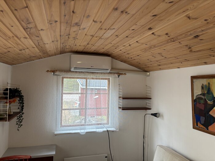 Rum med trätak, fönster med gardin, vägghängd hylla med växter och en installerad luftvärmepump ovanför fönstret.