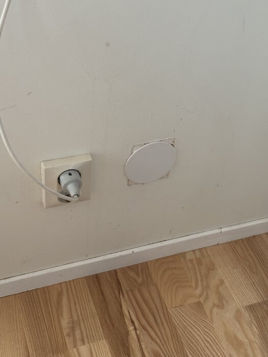 Vägg med elektriskt uttag och rund täckplatta, bredvid trägolv.