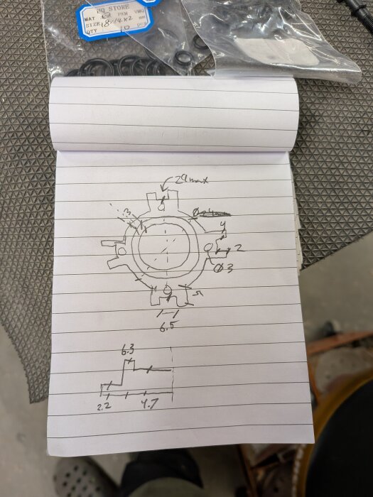 Blyertsskisser av en pistondämpare med mått på ett randigt anteckningsblock, omgiven av förpackningar med små O-ringar.