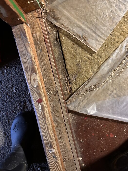 Närbild på ett golv bygge med träbalk, isolering av mineralull och plastfolie, en svart sko syns i nedre vänstra hörnet.