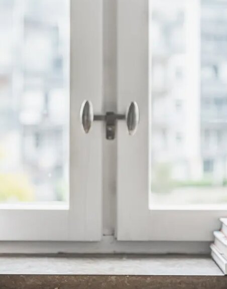 Inåtgående fönster i vitt med synliga handtag och del av karm, i fokus mot en suddig bakgrund av byggnader utanför.