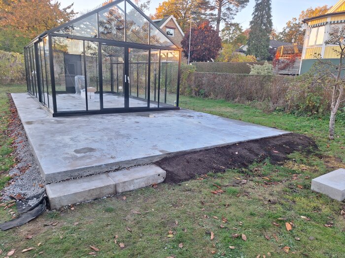 En glasbyggnad på en betongplatta i en trädgård med omgivande grönska och höstlöv.