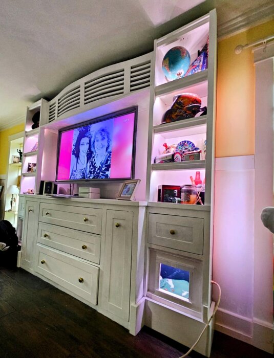 Väggmonterad enhet med två IKEA-hyllor och en byrå, anpassade med lister, MDF och färgad belysning, omger en TV i mitten.