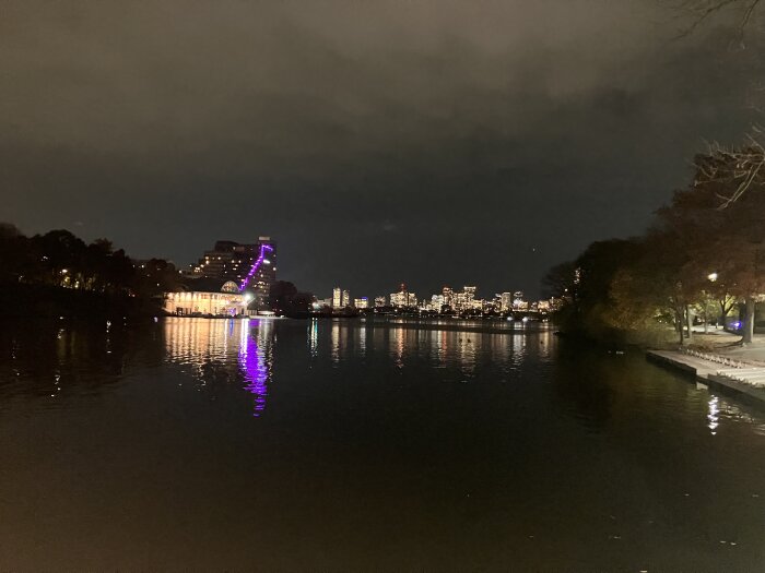 Kvällsbild av en stadssilhuett över vatten, med upplysta byggnader och träd samt reflektioner på ytan.