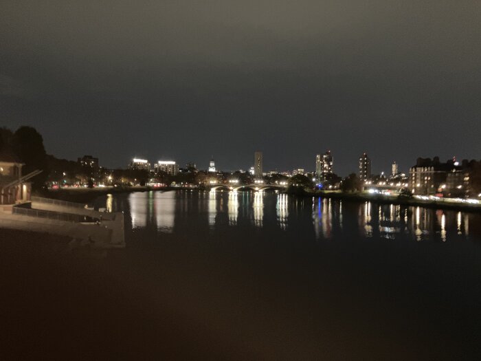 Stadssilhuett vid natt med upplysta byggnader och bro över en flod, reflektioner av ljus på vattnet.