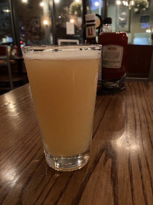 Ett glas med ljus öl stående på ett träbord i en restaurangmiljö med bakgrund av flaskor och svagt ljus.