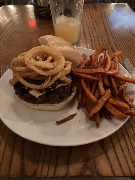 Hamburgare med lök, ost och sås serverad med sötpotatispommes på en tallrik. Halvt fylld glasdryck i bakgrunden på ett träbord.