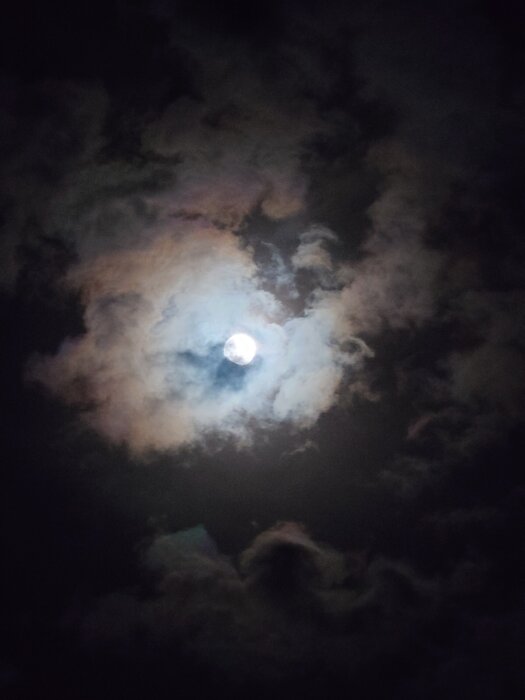 Månen lysande bakom mörka moln på nattlig himmel.