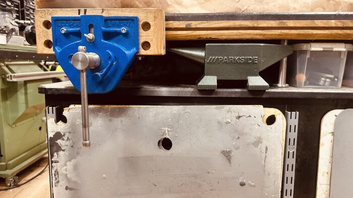 Gjutjärnsstädet på ett arbetsbord med blå skruvstycke, nära trä- och metallunderlag, med verktygslåda i bakgrunden.