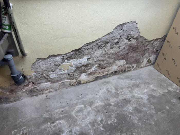 Avskalad puts på källarvägg med synliga stenblock och smågrus, nära ett avloppsrör i en hörna av ett rum.