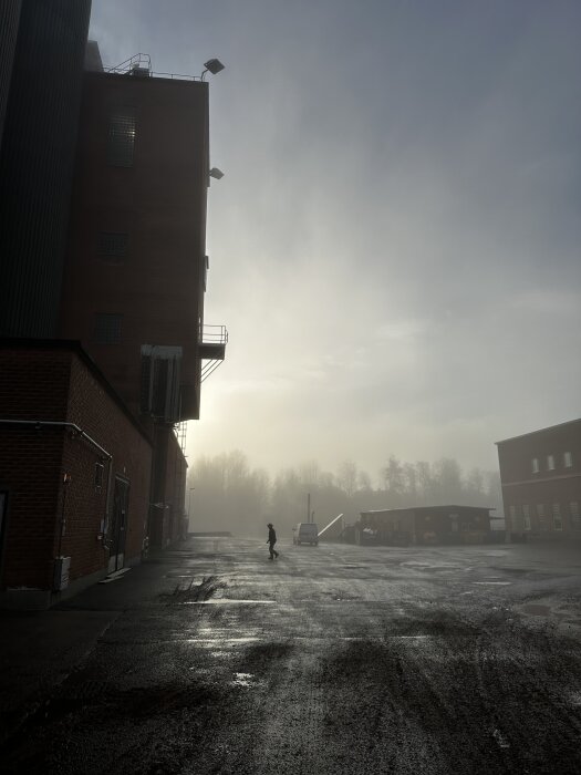 Dimma över en industribyggnad med en ensam person på en parkering och en bil i bakgrunden, tidig morgon eller sen eftermiddag.