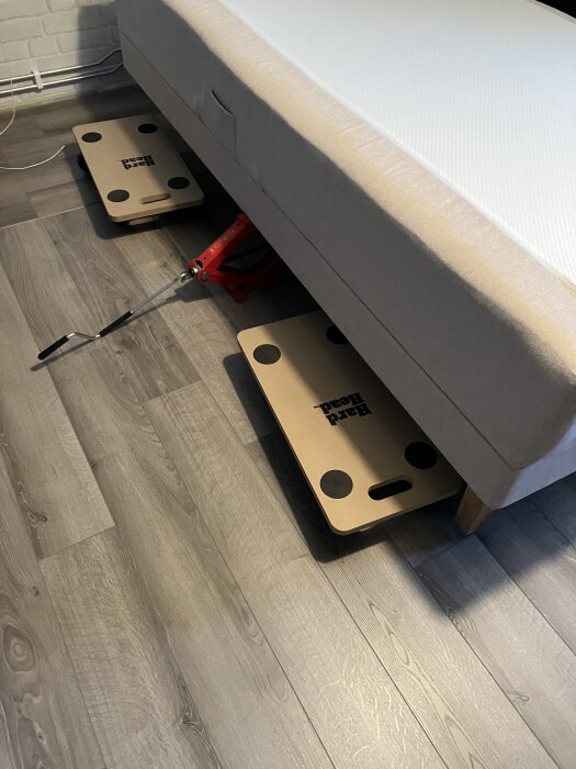 Tung säng på hjultransport och domkraft på ett trägolv, ben avskruvade för enklare förflyttning.