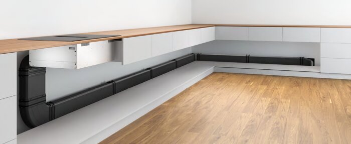 Inbyggd kanal bakom köksskåp med träbänkskiva och svart ventilation, sett längs hörnvägg med trägolv.