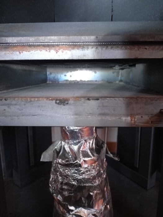 Inuti en kamin med borttagna askfat och synligt tilluftsrör i metall längst ner, omgiven av mörkt metallhölje.