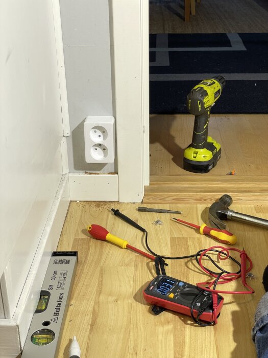 Inomhusmiljö med schneideruttag på väggen, en borrmaskin, skruvmejslar, hammare och multimeter på golvet.