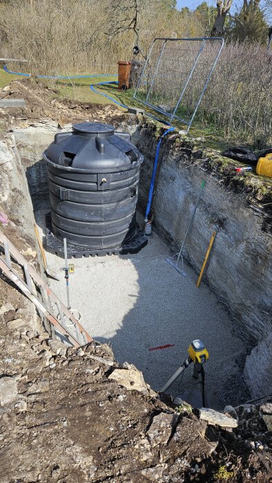 Svart avloppstank i grop med grusbotten, omgiven av olika verktyg och utrustning för installation.