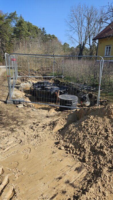Installation av avloppssystem med tankar och pumpbrunn i en grävd grop omgiven av staket, med en gul byggnad i bakgrunden.