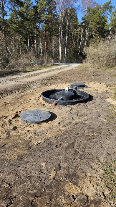 Bild av nedgrävda tankar och en pumpbrunn under installation på en öppen grusyta omgiven av träd.